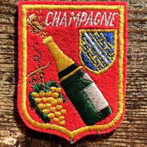 Pode incluir: Um emblema bordado vermelho com borda amarela apresentando uma garrafa de champanhe verde com gargalo dourado, um cacho de uvas amarelas com folhas verdes e um escudo azul com uma faixa diagonal branca. A palavra CHAMPAGNE está bordada em branco.