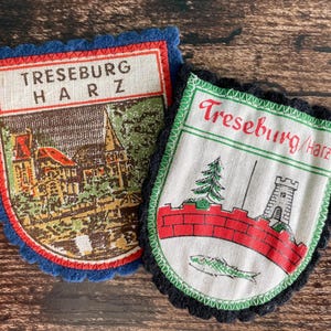May include: Two shield-shaped fabric patches with scalloped edges. The left patch has a blue border, the text TRESEBURG HARZ, and a detailed building illustration. The right patch has a black border, the text Treseburg/Harz, a red wall, a tree, and a fish.