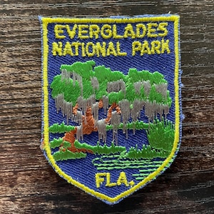 May include: Embroidered patch featuring the Everglades National Park. The patch has a blue background with a yellow border. The design includes a tree with green and grey foliage, and the text "EVERGLADES NATIONAL PARK FLA."