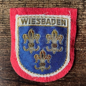 Pode incluir: Emblema bordado vermelho, branco e azul com a palavra "WIESBADEN". Apresenta três símbolos de flor-de-lis dourados sobre fundo azul, um design heráldico.