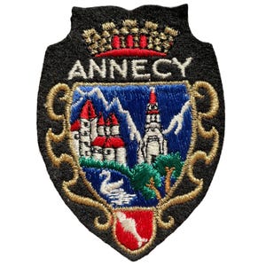 Op de afbeelding: Een schildvormige geborduurde patch met het woord ANNECY in wit garen. Het ontwerp bevat een gouden kroon, een blauw veld met witte bergen, een witte zwaan, groene bomen en een rode basis met een witte vorm, alles omlijnd met goudkleurig draad.