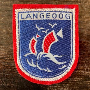 May include: A rectangular embroidered patch with a red felt border. The centre features a blue shield shape with the text LANGEOOG in white. Inside the shield are two stylised sailboats with red sails and white waves, with three white birds above.