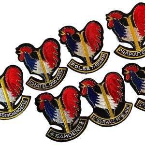 Op de afbeelding: Een verzameling van zeven geborduurde patches met een haanontwerp in rood, wit en blauw. Elke patch bevat een goudkleurige verticale balk en een gebogen banner aan de onderkant met tekst waaronder Besse-en-Chandesse, Chatel, Polset, Prapoutel, St. Gervais Mt. Blanc, Samoens en St. Nicolas.