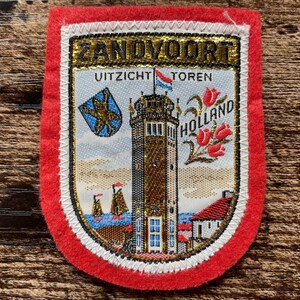 Peut inclure: Un écusson brodé rectangulaire avec une bordure en feutre rouge représentant une tour centrale, le texte ZANDVOORT, UITZICHT TOREN et HOLLAND, un écusson bleu avec un lion doré, deux voiliers sur l'eau et trois tulipes rouges sur fond crème.