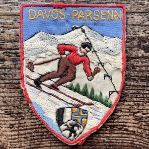 Peut inclure: Un écusson brodé en forme de bouclier portant le texte DAVOS-PARSENN en jaune sur un ciel bleu clair. Le motif représente un skieur en pull rouge et pantalon marron sur une pente enneigée avec des pins verts et un petit blason à la base.