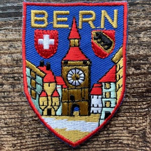 May include: A shield-shaped embroidered patch featuring the text BERN in gold letters. The design depicts a central clock tower with a red roof, flanked by buildings, a red shield with a white cross, and a red shield with a black bear emblem.