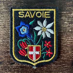 May include: Embroidered patch with the word "SAVOIE" in gold lettering. The patch features a shield with a red and white cross, and colorful floral designs including blue, white, and red flowers, with green stems and leaves, all outlined in gold.