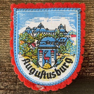 Op de afbeelding: Een schildvormige stoffen embleem met een rode geschulpte vilten rand. Het midden toont een gedrukte illustratie van een kasteelcomplex, bomen en een klein wapenschild met een rood hert. De tekst Augustusburg staat onderaan in het zwart.