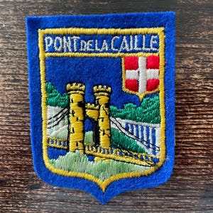 Può includere: Una toppa ricamata blu con la scritta PONT DE LA CAILLE in filo bianco. Il disegno raffigura due torri in pietra gialla collegate da un ponte, elementi paesaggistici verdi e uno scudo con una croce rossa e bianca su sfondo blu.