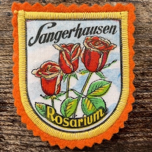 Peut inclure: Un écusson brodé en forme de bouclier avec une bordure en feutre orange festonnée et un contour jaune. Le centre présente un fond blanc avec trois roses rouges, des feuilles vertes et le texte noir Sangerhausen et Rosarium.