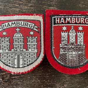 May include: Two red and white embroidered patches featuring the word "HAMBURG" above a castle design. The patches are shield-shaped, possibly souvenirs or emblems, with a white border.