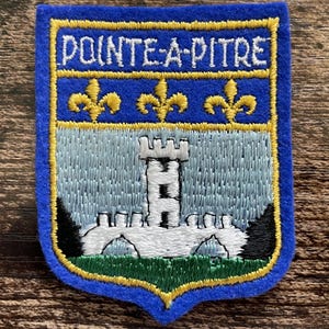 Könnte beinhalten: Ein schildförmiger, bestickter Aufnäher mit blauem Rand und gelber Umrandung. Der obere Teil zeigt den Text POINTE-A-PITRE in Weiß auf blauem Grund mit drei gelben Lilien. Der untere Teil stellt eine weiße Burg auf hellblauem Hintergrund dar.