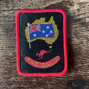 May include: A rectangular embroidered patch with a black background and a bright red border. It features a gold-colored map of Australia, a blue Australian flag, a red kangaroo silhouette, and the word AUSTRALIA in red capital letters.
