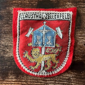 May include: A red and white embroidered patch with the text "CLAUSTHAL-ZELLERFELD" at the top. The patch features a blue and white emblem with a cross and two figures, and a gold lion at the bottom.
