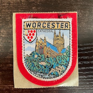 Może przedstawiać: Tkana naszywka z czerwoną obwódką i białym przeszyciem, przedstawiająca katedrę w Worcester. U góry znajduje się napis "WORCESTER CATHEDRAL". Obraz przedstawia katedrę z niebieskimi i złotymi detalami.
