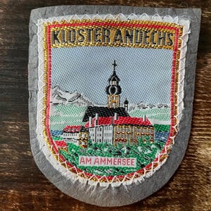 Può includere: Una toppa ricamata a forma di scudo con cielo azzurro, una chiesa con un campanile scuro, tetti rossi e un paesaggio verde. Il testo KLOSTER ANDECHS è in alto e AM AMMERSEE in basso. La toppa ha un bordo in feltro grigio.