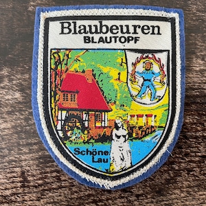Peut inclure: Un écusson brodé en forme de bouclier avec le texte Blaubeuren Blautopf et Schöne Lau. Le motif représente un bâtiment au toit rouge avec une roue à aubes, un blason blanc avec une figure bleue et une statue blanche sur un paysage vert.