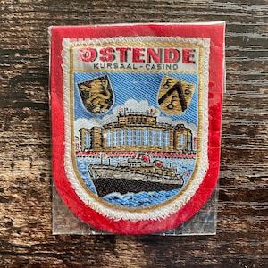 Może przedstawiać: Haftowana naszywka z napisem "OSTENDE KURSAAL-CASINO" przedstawiająca statek, budynek i dwa herby. Naszywka ma czerwoną obwódkę i jest w przezroczystym plastikowym rękawie.