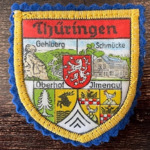 Op de afbeelding: Een schildvormige stoffen patch met een blauwe geschulpte rand en een gele gestikte boord. De bovenste rode baan bevat de tekst Thüringen. Daaronder bevinden zich vier kwadranten met Gehlberg, Schmücke, Oberhof en Ilmenau en een centraal rood schild.