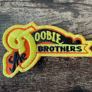 Può includere: Toppa ricamata con la scritta "The Doobie Brothers". La toppa presenta una combinazione di colori giallo, arancione e nero. Il testo è in un font stilizzato, con un bordo giallo.