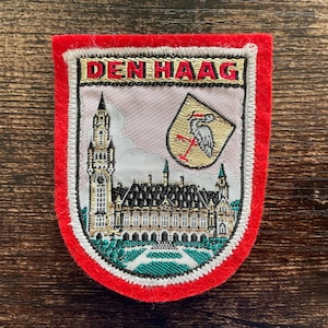 Op de afbeelding: Geborduurde patch met een rode rand met bovenaan de tekst "DEN HAAG". De patch toont een gedetailleerde illustratie van een gebouw met een klokkentoren en een wapenschild met een ooievaar. De patch ligt op een houten oppervlak.