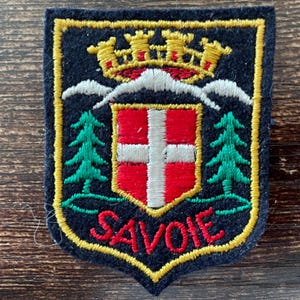 Peut inclure: Un écusson brodé en forme de blason sur fond bleu marine. Il présente une couronne dorée au-dessus de montagnes blanches, deux sapins verts et un écusson central rouge avec une croix blanche. Le mot SAVOIE est brodé en rouge en bas.