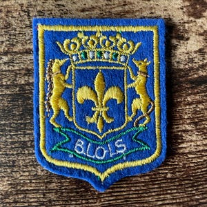 May include: Blue embroidered patch with a gold fleur-de-lis, two gold lions, and a gold crown on a shield. The patch has the word "BLOIS" in green and white. The patch is bordered in gold and has a blue background.