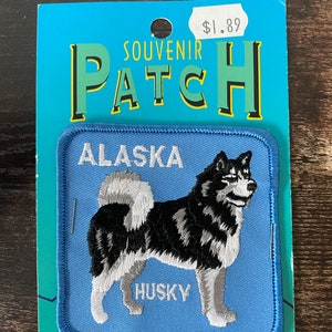 Könnte beinhalten: Ein Souvenir-Patch mit einem schwarz-weiß-grauen Husky-Hund auf hellblauem Hintergrund. Der Patch ist mit den Worten "ALASKA" und "HUSKY" bestickt. Der Patch ist an einer blauen Karte mit dem Wort "SOUVENIR PATCH" befestigt.