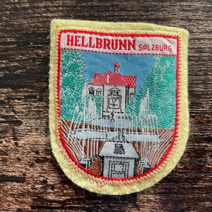May include: A rectangular embroidered patch with a pale yellow border featuring a scene of Hellbrunn Palace in Salzburg. The design includes a white building with a red roof, green trees, and water fountains, with the text HELLBRUNN SALZBURG.
