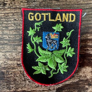 May include: A shield-shaped embroidered patch with a black background and a red border. The text GOTLAND is stitched in yellow at the top. A central blue crest features a white ram, surrounded by green embroidered vine and leaf motifs.