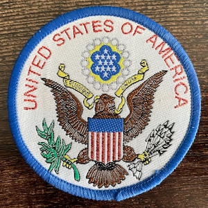 Puede incluir: Un parche bordado circular que presenta el Gran Sello de los Estados Unidos. El diseño incluye un águila marrón con un escudo, sosteniendo una rama de olivo y flechas, rodeado por el texto UNITED STATES OF AMERICA en rojo sobre fondo blanco.
