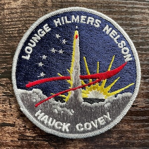 May include: Embroidered patch with a space theme. The patch depicts a rocket launching, set against a blue backdrop with white stars. The names "LOUNGE HILMERS NELSON" and "HAUCK COVEY" are embroidered in white. Red and yellow details.