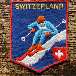 Op de afbeelding: Een rechthoekige geborduurde patch met een blauwe achtergrond en een rode rand. De tekst SWITZERLAND is in het geel bovenaan geborduurd. Het toont een skiër in een blauw pak, oranje muts en handschoenen op een witte helling met een rode Zwitserse vlag.