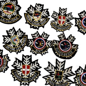 May include: A selection of embroidered patches with black and white snowflake patterns. Each patch displays a ski resort name, including "La Clusaz" and "Mont-Louis 1650m", with vibrant illustrations of skiers.