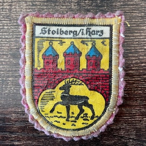 May include: A vintage patch with a shield design featuring the text "Stolberg/i.Harz". The patch has a yellow background with red castle illustrations and a black deer. The border is pink.