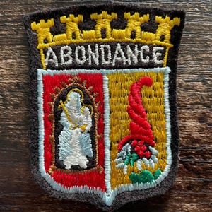 May include: A shield-shaped embroidered patch with a black background featuring a yellow mural crown above the text ABONDANCE. The shield is divided into a red section with a white figure and a yellow section with a red cornucopia.