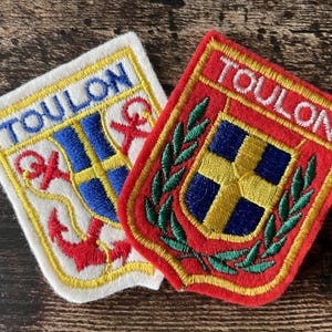May include: Two embroidered patches featuring the text TOULON. One patch has a white background with a blue and yellow shield, red anchors, and yellow trim. The other patch has a red background with a blue and yellow shield, green laurel leaves, and red trim.