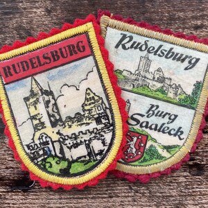 Op de afbeelding: Twee schildvormige stoffen patches met geborduurde randen. De linker patch heeft een rode banner met de tekst RUDELSBURG boven een kasteelillustratie. De rechter patch toont de tekst Rudelsburg en Burg Saaleck met een kasteel en een klein rood schild.