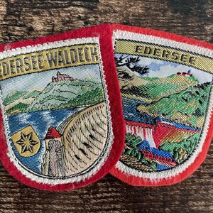 Puede incluir: Dos parches bordados en forma de escudo con bordes de fieltro rojo. El parche izquierdo presenta el texto EDERSEE WALDECK, un castillo en una colina, una presa y un escudo dorado con una estrella. El parche derecho muestra el texto EDERSEE.