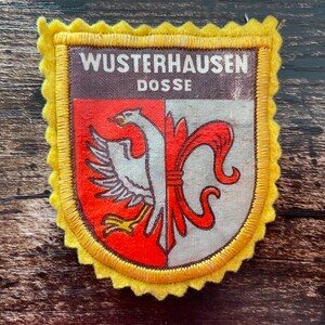Op de afbeelding: Een schildvormige stoffen patch met een gele geschulpte rand. Het bovenste gedeelte is donkerbruin met de witte tekst WUSTERHAUSEN DOSSE. Het onderste gedeelte is verdeeld in rode en witte helften, met een witte adelaar en een rode fleur-de-lis.