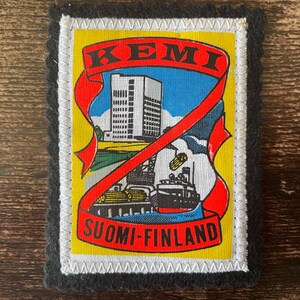 May include: A rectangular embroidered patch featuring a yellow background with a red ribbon banner containing the text KEMI and SUOMI-FINLAND. The center depicts a white building, a ship, and industrial elements in blue, black, and white.