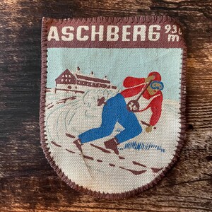 Op de afbeelding: Een rechthoekige stoffen patch met afgeronde onderhoeken met een skiër in een rood jack en blauwe broek op een besneeuwde helling. De tekst ASCHBERG 936 m staat bovenaan. De patch heeft een bruine rand met zichtbare gestikte randen.