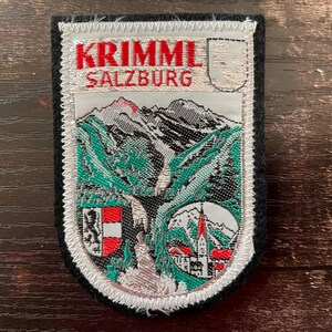 May include: Embroidered patch with the text "KRIMML SALZBURG" in red. The patch shows a mountain landscape with green and white details, a waterfall, and a small town with a church. It has a black border.