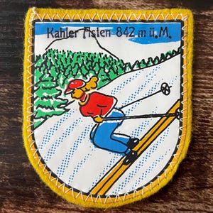 Op de afbeelding: Een schildvormige geborduurde patch met een gele rand met een afbeelding van een skiër in een rood jack en blauwe broek op een besneeuwde helling. De tekst "Kahler Asten 842 m ü.M." staat bovenaan boven een groen bos en bergsilhouet.