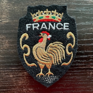Op de afbeelding: Geborduurde patch in schildvorm met het woord "FRANCE" in wit. Een gouden haan met rode kam is gecentreerd onder de tekst, met een kroon erboven. De patch is zwart met gouden en rode accenten.