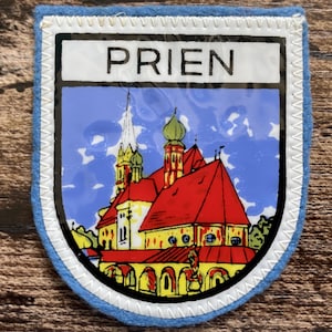 May include: A shield-shaped embroidered patch with a light blue felt border. The centre features a glossy illustration of a church with red roofs and yellow walls against a blue sky. The word PRIEN is printed in black capital letters at the top.