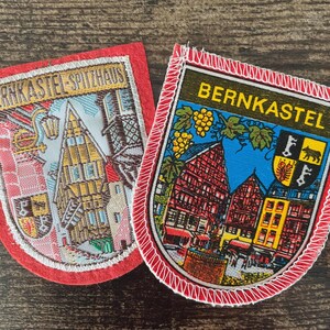 Könnte beinhalten: Zwei gestickte Aufnäher mit rotem Rand. Der linke Aufnäher zeigt ein Gebäude und den Text BERNKASTEL-SPITZHAUS. Der rechte Aufnäher zeigt Fachwerkhäuser, Weintrauben, ein Wappen und den Text BERNKASTEL in gelben Buchstaben.
