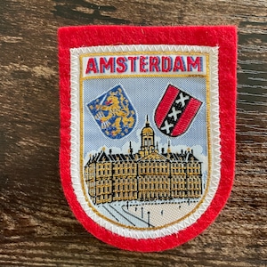 May include: A red felt patch featuring an embroidered design of the Amsterdam Royal Palace. The patch includes the text AMSTERDAM in red letters, a blue shield with a gold lion, and a red shield with three white crosses on a light blue background.