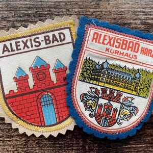Könnte beinhalten: Zwei bestickte Stoffaufnäher mit gezackten Rändern. Der linke Aufnäher ist beige mit einer roten Burg und dem Text ALEXIS-BAD. Der rechte Aufnäher ist blau mit dem Text ALEXISBAD HARZ KURHAUS, einer Gebäudeabbildung und einem Wappen.