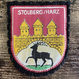 Könnte beinhalten: Ein Stoffaufnäher mit schwarzem Bogenrand und einem rot-gelben Wappenmotiv. Der Text STOLBERG/HARZ ist oben aufgedruckt. Die Grafik zeigt eine gelbe Steinmauer mit drei Türmen über der schwarzen Silhouette eines Hirsches auf hellem Grund.